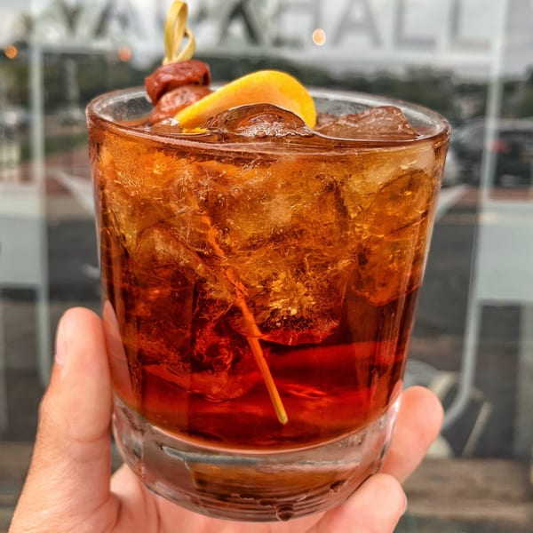 A hand holds a Vauxhall cocktail with ice, an orange slice, and cherries, against a blurred background.