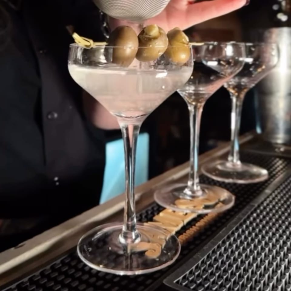 A bartender garnishes a martini with three olives on a skewer, with two more glasses in the background.