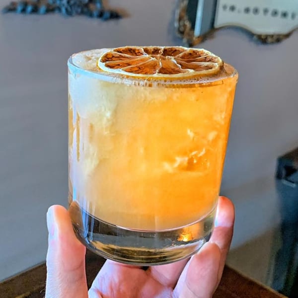 A hand holds a short glass of orange cocktail with a dried citrus slice garnish.