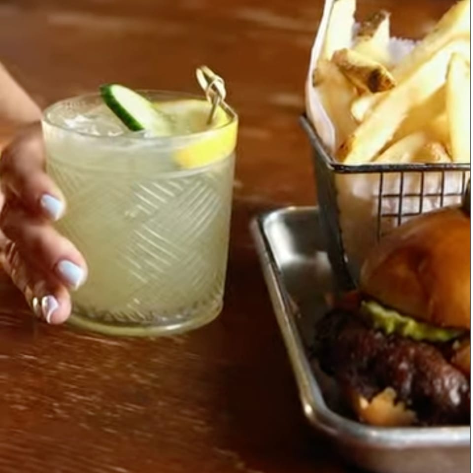 A hand holding a cocktail with cucumber and lemon, next to a burger and fries.