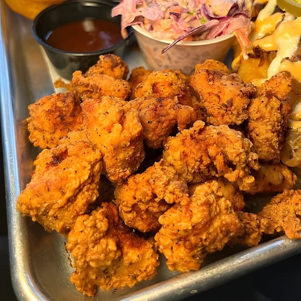 Crispy fried chicken bites on a metal tray with coleslaw, sauce, and cheesy fries.