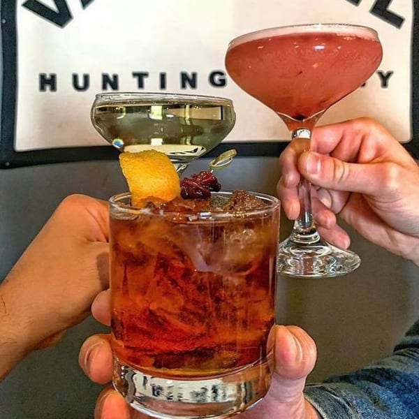 Three hands holding three different cocktails, including a red one and an amber one with orange peel.
