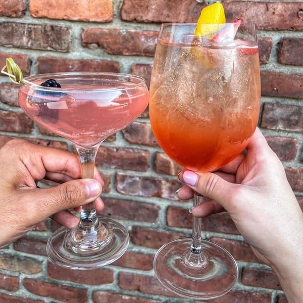 Two hands holding pink cocktails with fruit garnishes against a brick wall.