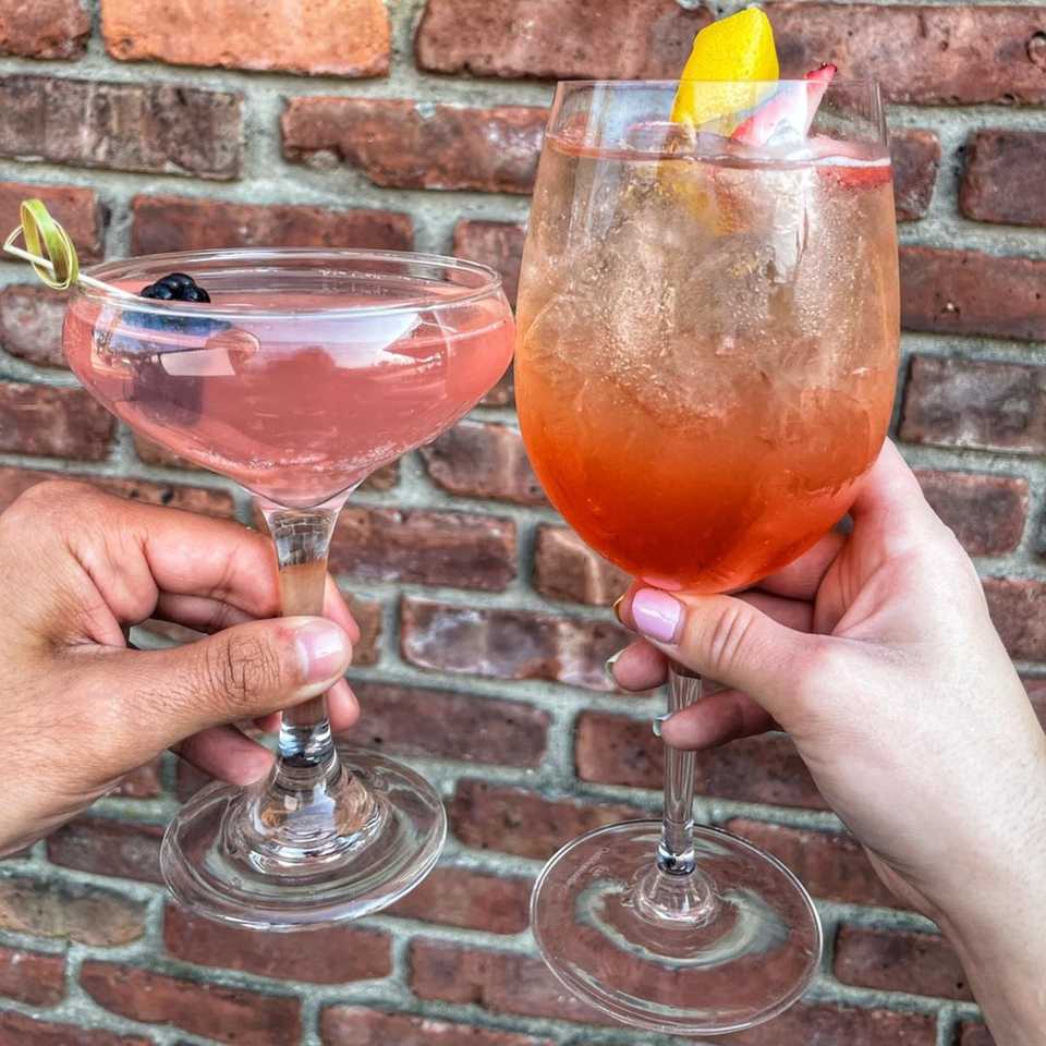Two hands holding pink cocktails with fruit garnishes against a brick wall.