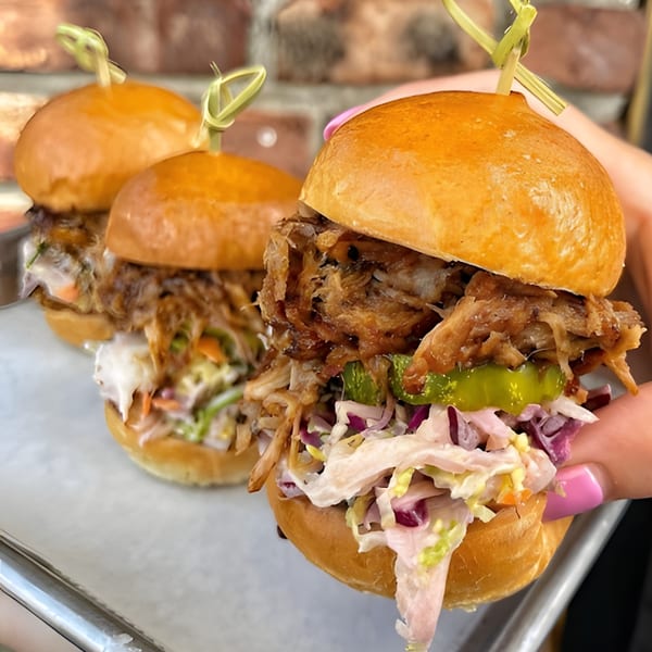 Three pulled pork sliders with coleslaw and pickles on a tray, one held by a hand.