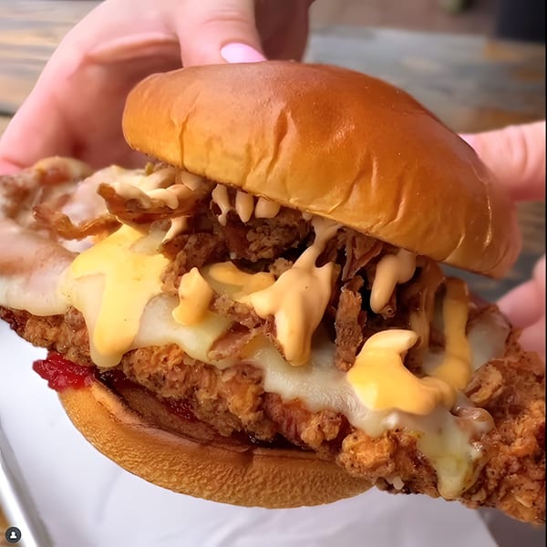 Hands holding a fried chicken sandwich with melted cheese, crispy onions, and sauce on a brioche bun.