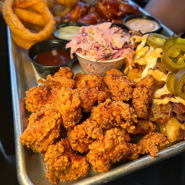 A metal tray filled with fried chicken bites, onion rings, coleslaw, and loaded fries with jalapeños.